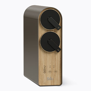 A modern kitchen appliance with a wood finish, featuring two large black dials on the front and labeled buttons below. The device has a sleek, rectangular design and the brand name Brio on the front.